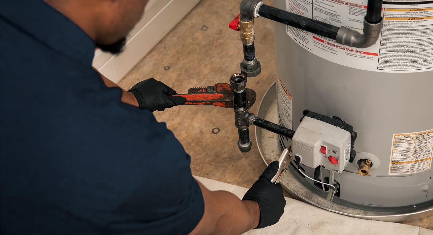 A Gold Medal plumber wearing black gloves uses a pipe wrench to repair the gas line and control valve assembly on a residential water heater tank.