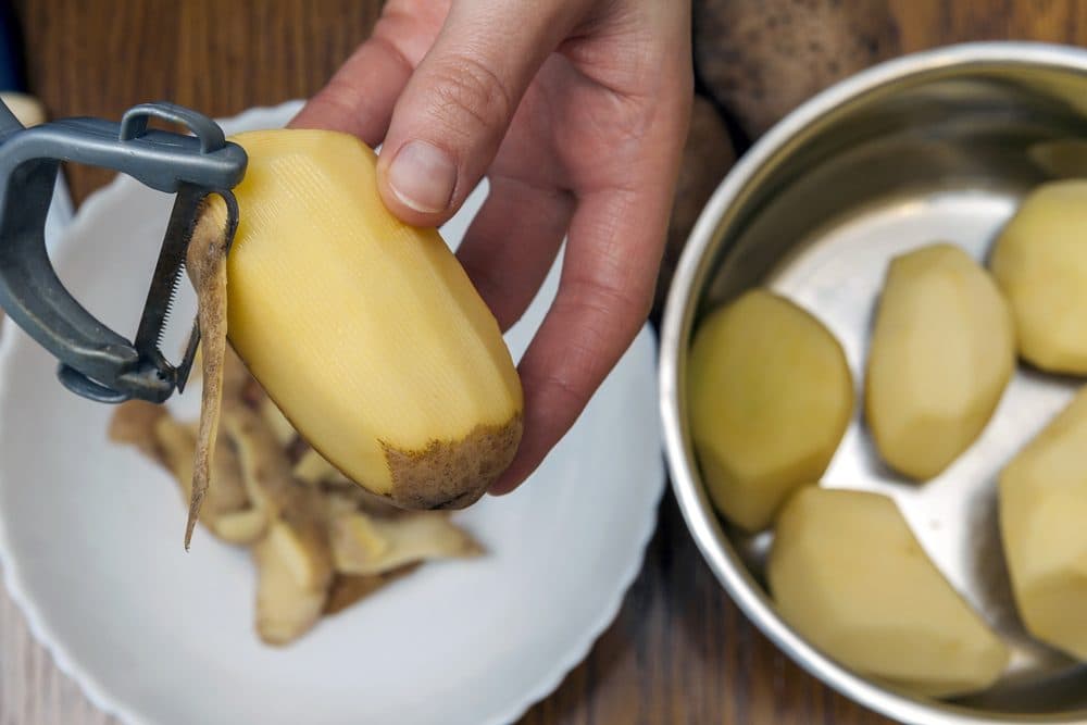 A potato is being peeled with a potato peeler with peels falling onto a plate with a pot of peeled potatoes in the background