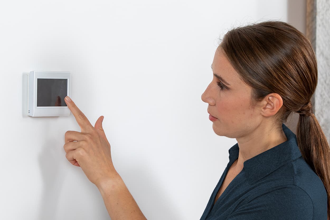 Side profile of a woman interacting with a digital programmable thermostat installed on a white wall.