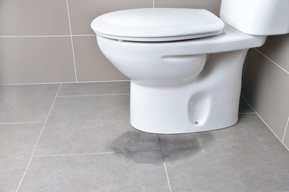 A toilet inside of a gray tiled bathroom shows visible signs of leaking on the floor