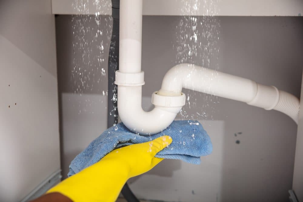 A hand in a yellow glove attempts to catch water leaking out of a pipe with a blue towel