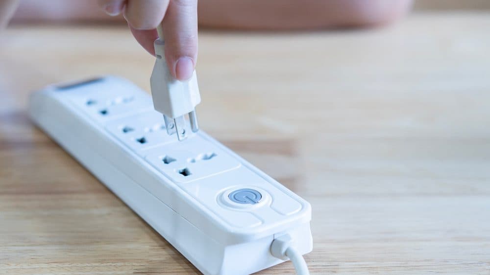 A hand is seen plugging a power cord into a power strip resting on the floor