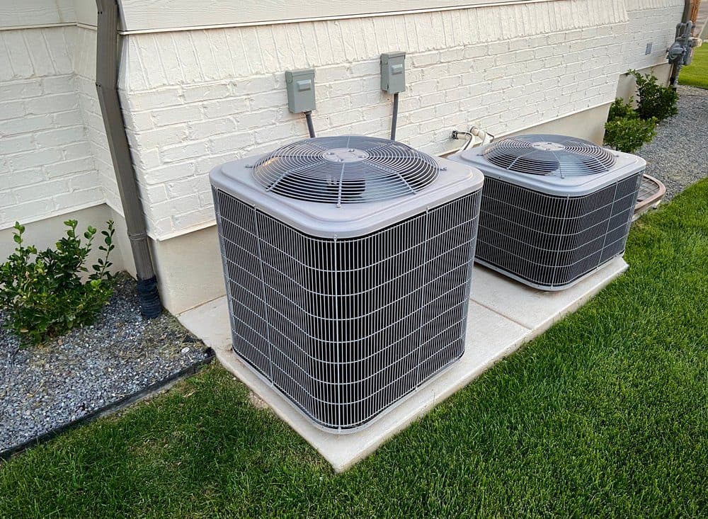 Two outdoor HVAC units sitting next to a beige house and a manicured lawn